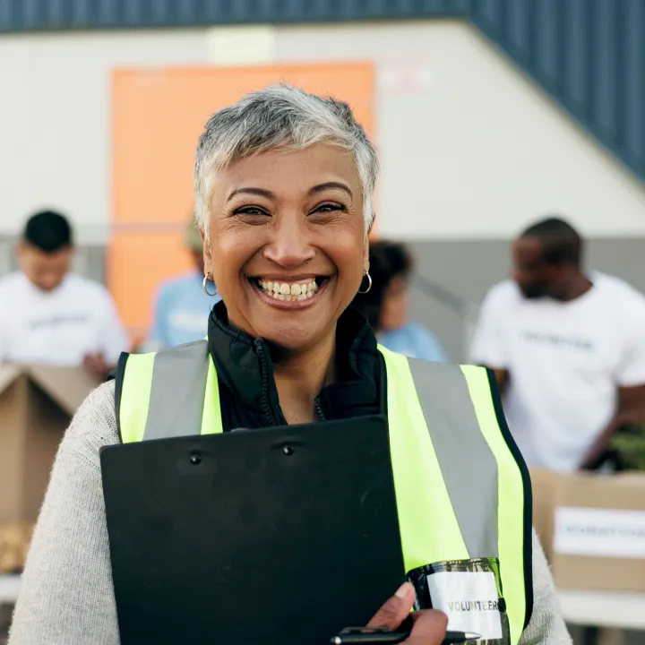 Smiling woman volunteer in reflective vest holds clipboard; others sort donation boxes.