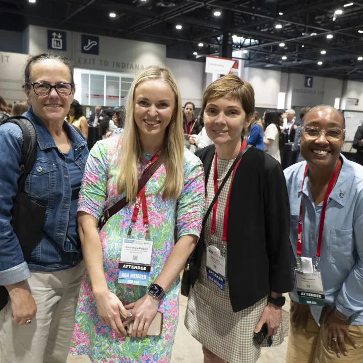 Four smiling women with lanyards pose at a conference.
