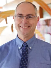 Smiling man with glasses, blue button-up shirt, and patterned tie.