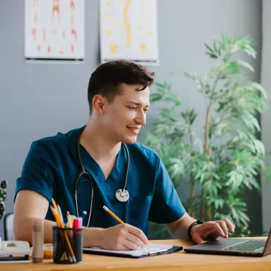 Smiling male health care worker in blue scrubs and stethoscope around neck looking at laptop computer and taking notes