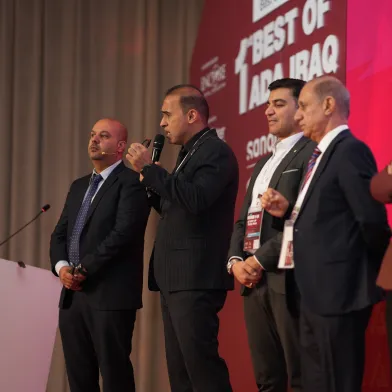 Five men on a stage; one speaks into a microphone. Banner partially reads "BEST OF ADA IRAQ".
