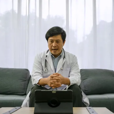 Male doctor in lab coat sits on sofa, engaging in a virtual consultation via tablet.