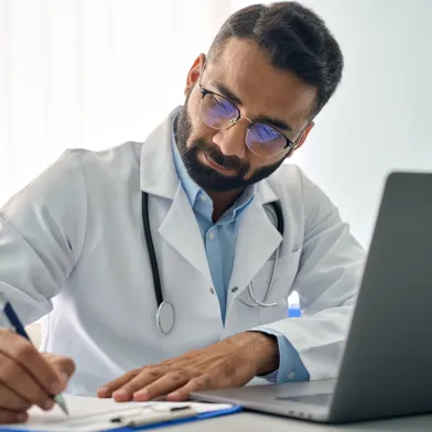 Focused male doctor in lab coat writes on clipboard next to a laptop.