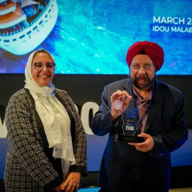 Smiling woman and man in a turban on stage; man holds a clear award plaque.