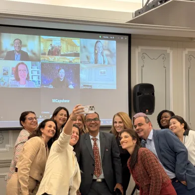 Smiling group poses for a selfie, a virtual meeting displayed on screen behind them.