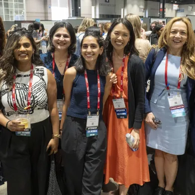 Seven smiling women, wearing lanyards, pose together at a conference.