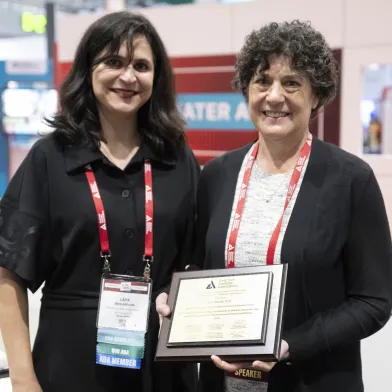 Two smiling women, one holding an award plaque, at a conference.
