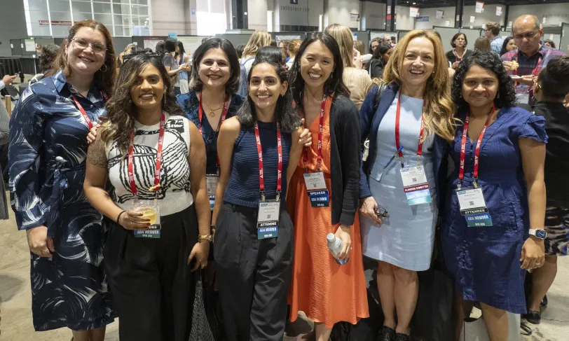 Seven smiling women, wearing lanyards, pose together at a conference.