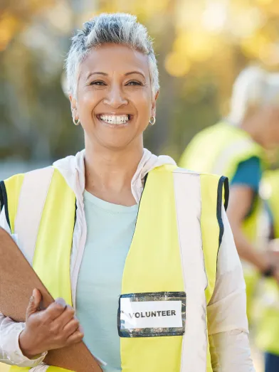 Smiling woman volunteer in a yellow vest holding a clipboard, with a group outdoors.
