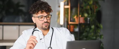 Photograph: Male doctor in white coat with stethoscope holds a medicine bottle, looking at a laptop.