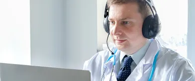 Doctor in white coat and headphones uses laptop for telehealth consultation.