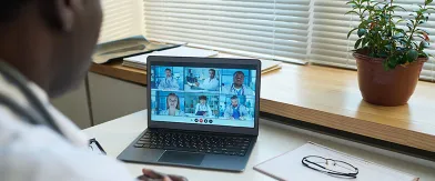 Doctor in white coat on video call with 6 other medical professionals on laptop.