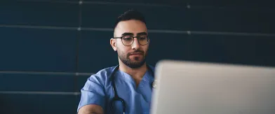 Male healthcare professional in blue scrubs, glasses, and stethoscope looks at laptop.