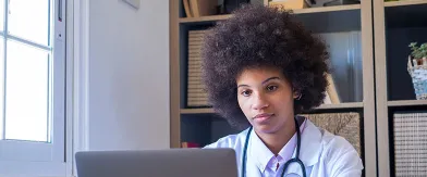 Young Black woman in lab coat with stethoscope, looking at a laptop.