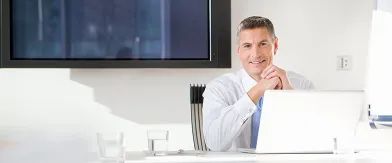 Professional man smiling at desk with laptop, hands clasped, in an office.