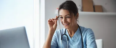 Smiling medical professional in scrubs with stethoscope looking at laptop.