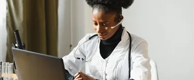 Focused Black doctor in lab coat, headset, viewing laptop screen.