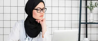 Female doctor in hijab, glasses, and stethoscope looks thoughtfully at laptop.