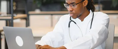 Focused Black doctor in a lab coat and stethoscope using a laptop.