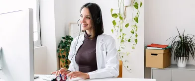 Woman in white shirt and headset smiles at computer screen, working remotely.